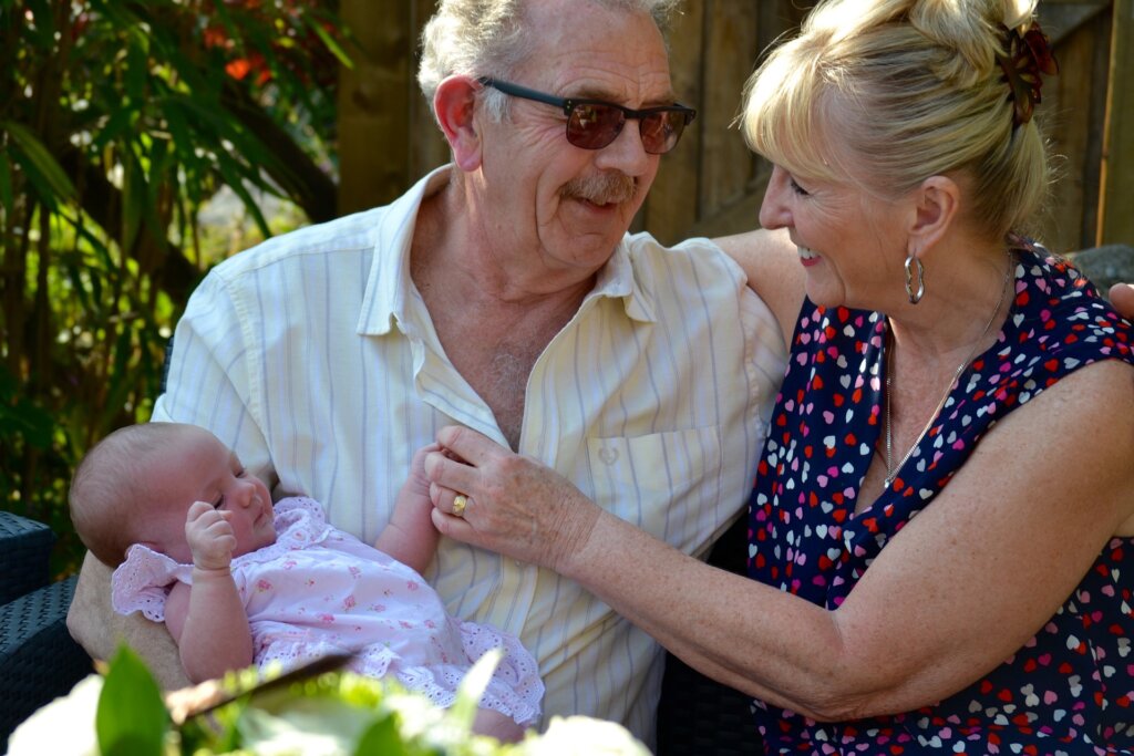 Image of a Caucasian couple who is likely in their 70's. They appear to be seated outside at a table enjoying a meal. The man is wearing sunglasses and a nice short-sleeved dress shirt. The woman is wearing a print blouse or dress and has her beautiful blond hair pinned up on her head. The man is holding a beautiful baby girl who is wearing a printed pink onesie. The image is shown in conjunction with our Braintree estate planning law firm's blog post about Medicare premiums and long term care planning.
