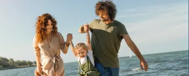 This image shows a happy family walking along the beach. A woman with curly reddish hair is holding up the arms of a blond-haired girl who appears to be aged four or other. To the right of the little girl a very tall man is holding up the girl's other arm. The couple appears to be walking along the beach while swinging their daughter as they walk along the beach. The image is shown to support the fact our Braintree MA law firm can help families with long term planning, including creating a last will and testament.