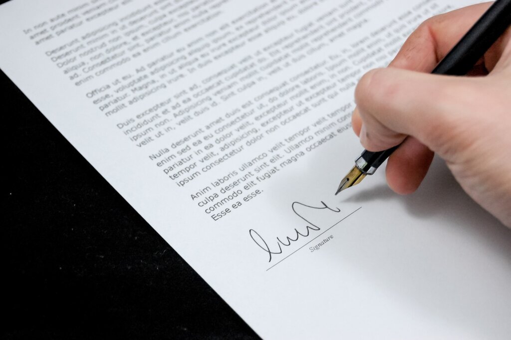 This is the image of a hand hovering over a printed document. The hand is holding a black fountain pen and signing a document. In this case, the image being shown is related to our estate planning firm's blog post about the difference between a living will and DNR (Do Not Resuscitate) document.
