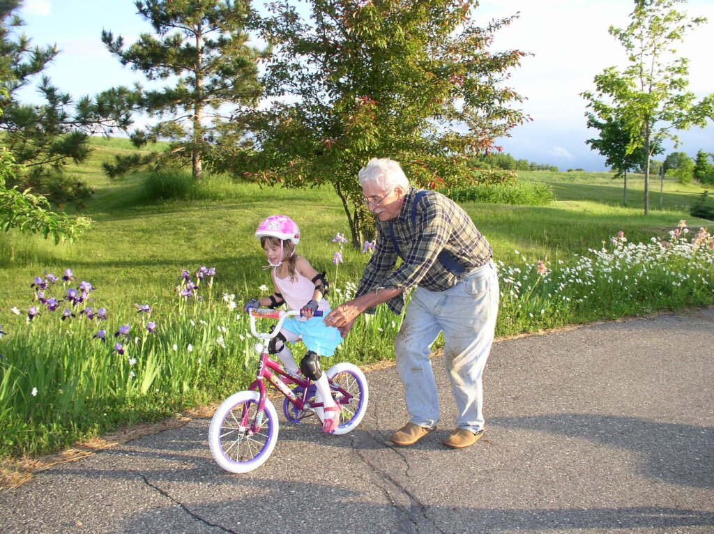 Image of a paved road in the middle of a beautiful park or residential complex that has pine and other trees and lots of pink, white and purple wildflowers growing next to the paved road. A grandfather who is wearing glasses is pushing his granddaughter on her pink bike. The granddaughter is about 4 or 5 and is wearing a bike helmet with pink flowers and has long blond hair. This image is shown in conjunction with our Braintree estate planning firm's blog post about long term care insurance and long term care planning.
