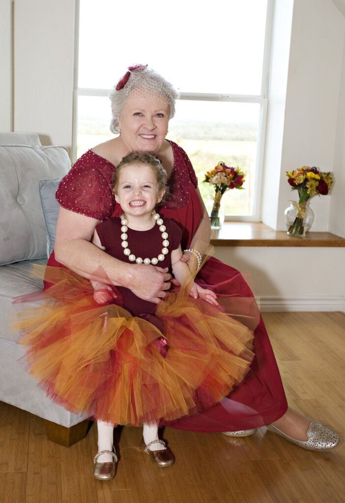 Image of a beautiful older woman seated on a light-colored couch with her granddaughter standing up against her. Both are smiling and facing the camera. Both are wearing maroon. The woman has on a little hat with white netting and maroon flowers that covers the top of her short white hair. The little girl has a maroon top and orange and maroon ballet tutu and his wearing a string of pearls with big beads. The image is shown related to our Braintree estate planning law firm's blog post about Medicare and long term care planning.
