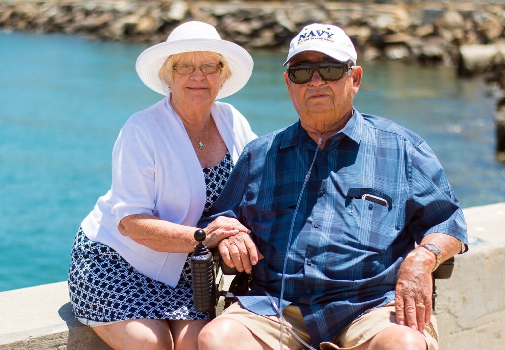 This in an image of a couple who appears to be in their seventies or eighties. They are sitting on or near a wall overlooking a beautiful blue body of water. The woman is wearing a white hat, a long, modest one-piece blue & white bathing suit and a white cardigan sweater. She is holding the left arm of a man seated in a wheelchair who appears to be attached to portable oxygen. He is wearing a baseball cap that says "Navy" and a nice short-sleeved plaid dress shirt of summery blues and tan shorts. The image is shown related to our estate planning firm's blog post about long term care planning and what happens to one spouse if another qualifies for MassHealth/Medicaid.