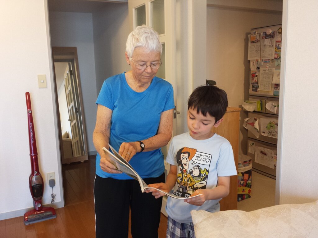 Image of a grandmother with her grandson. The grandmother has short white hair and glasses and is wearing a bright royal blue t-shirt and is holding the right-hand side of a book. Her grandson is standing to the right of her holding the other half of the book. He has short black hair and is wearing a t-shirt that includes characters from "Toy Story."  They appear to be standing in a family room just off the kitchen. The image is shown related to our Braintree estate planning firm's blog post about irrevocable trusts and asset protection.
