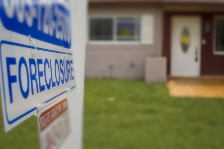 This is an image shows a foreclosure sign in high resolution in the forefront, and a home in the background in blurred/low resolution. It is shared in keeping with our blog post about how seniors can lose their equity in their home, plus miss out on transferring wealth to the next generation, by not paying their property taxes.
