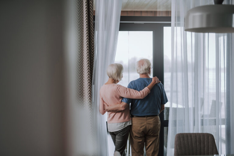 View of senior woman and man looking out window of home and embracing. This image is in keeping with our elder law firm's blog post about how to avoid foreclosure with a repayment plan for reverse mortgages.
