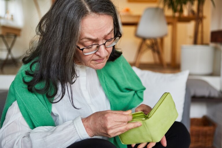 Senior-woman-at-home-looking-worriedly-into-her-empty-wallet. This image is shown in conjunction with our estate planning firm's blog post about how selling your life insurance may help you cover expenses, such as long-term care.