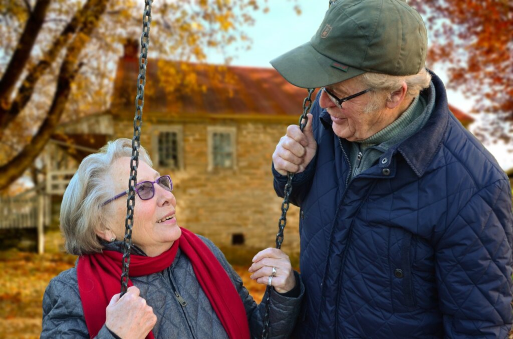 Image shows an elderly woman sitting on the swing of the backyard of a yellow house who is surrounded by an elderly gentleman who is standing over her and talking to her and who appears to be her husband and both the man and woman are smiling and happy.