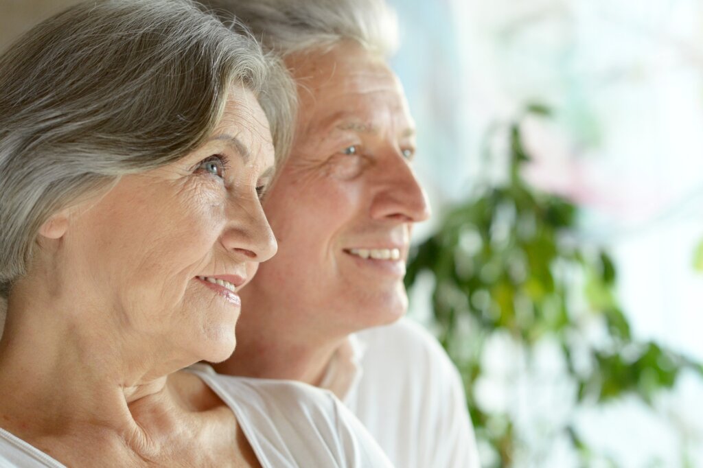 Image shows the shoulders and above of an attractive older couple sitting very close together -- a woman with gray and white short hair wearing lip stick is on the left and a man with gray and white hair is sitting on the right.