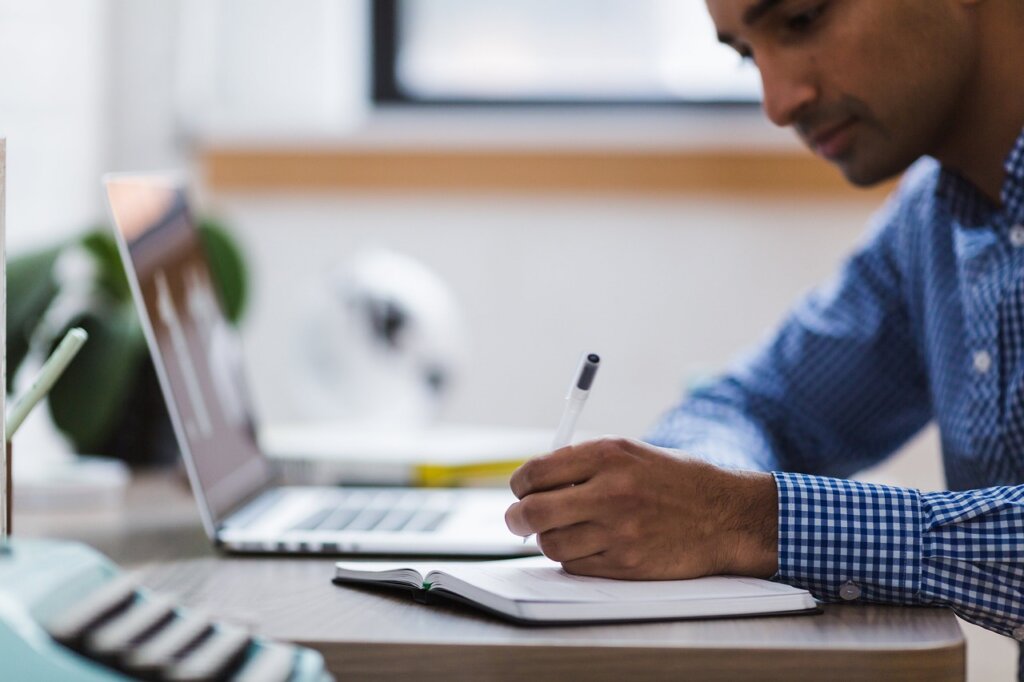 This rectangular image shows a sideview of a laptop computer and a dark-skinned handsome young man wearing a blue and white checkered shirt sitting in front of it taking notes in a notebook.