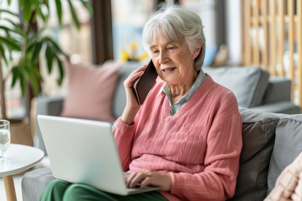 This is the image of a pretty older woman who has short gray hair and who is wearing a pink cable-knit v-neck sweater over a printed blouse and who is sitting on her couch with her hand on an open laptop computer while also talking at the same time on a mobile phone.