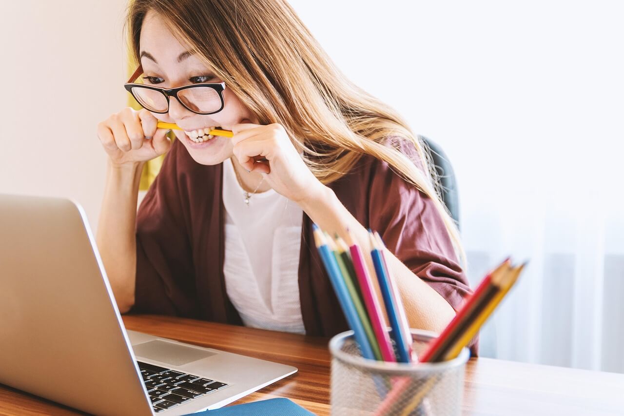 image of a middle-aged asian woman with long hair wearing glasses biting a yellow pencil between her teeth while she stares at her laptop computer screen